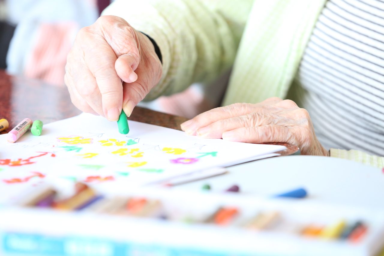 gallery-01 Faceless elderly woman with crooked fingers drawing with crayons at table in nursing home